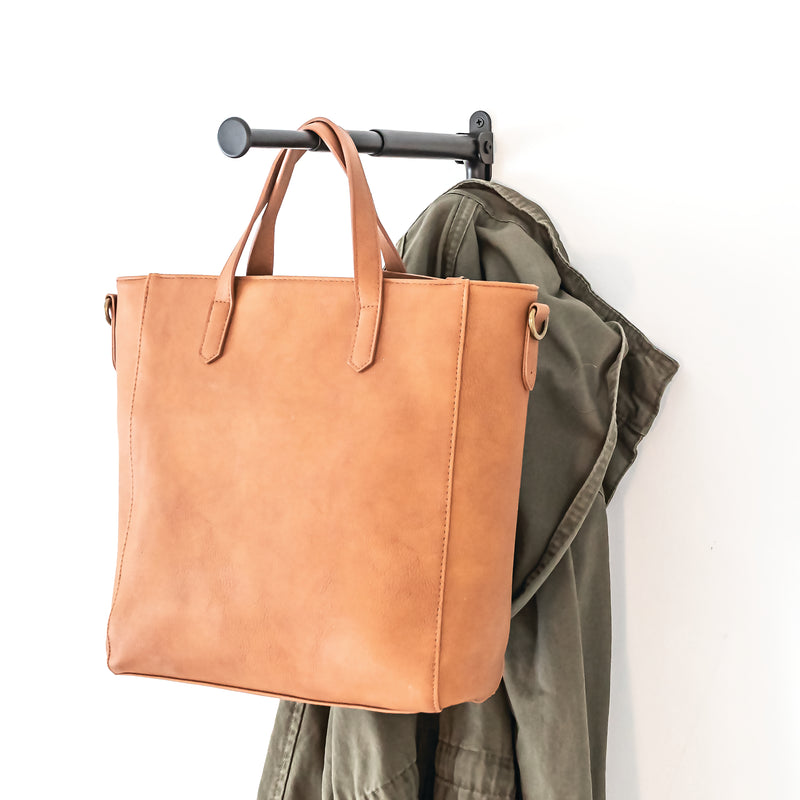 Brown leather tote bag hanging on a rack with a green jacket.