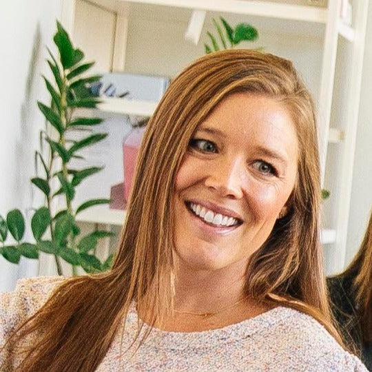 Woman with long light brown hair smiling in a casual setting with plants and shelves in the background