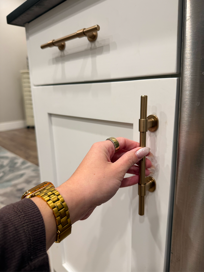 Hand with gold watch opening a white cabinet door with a gold handle.